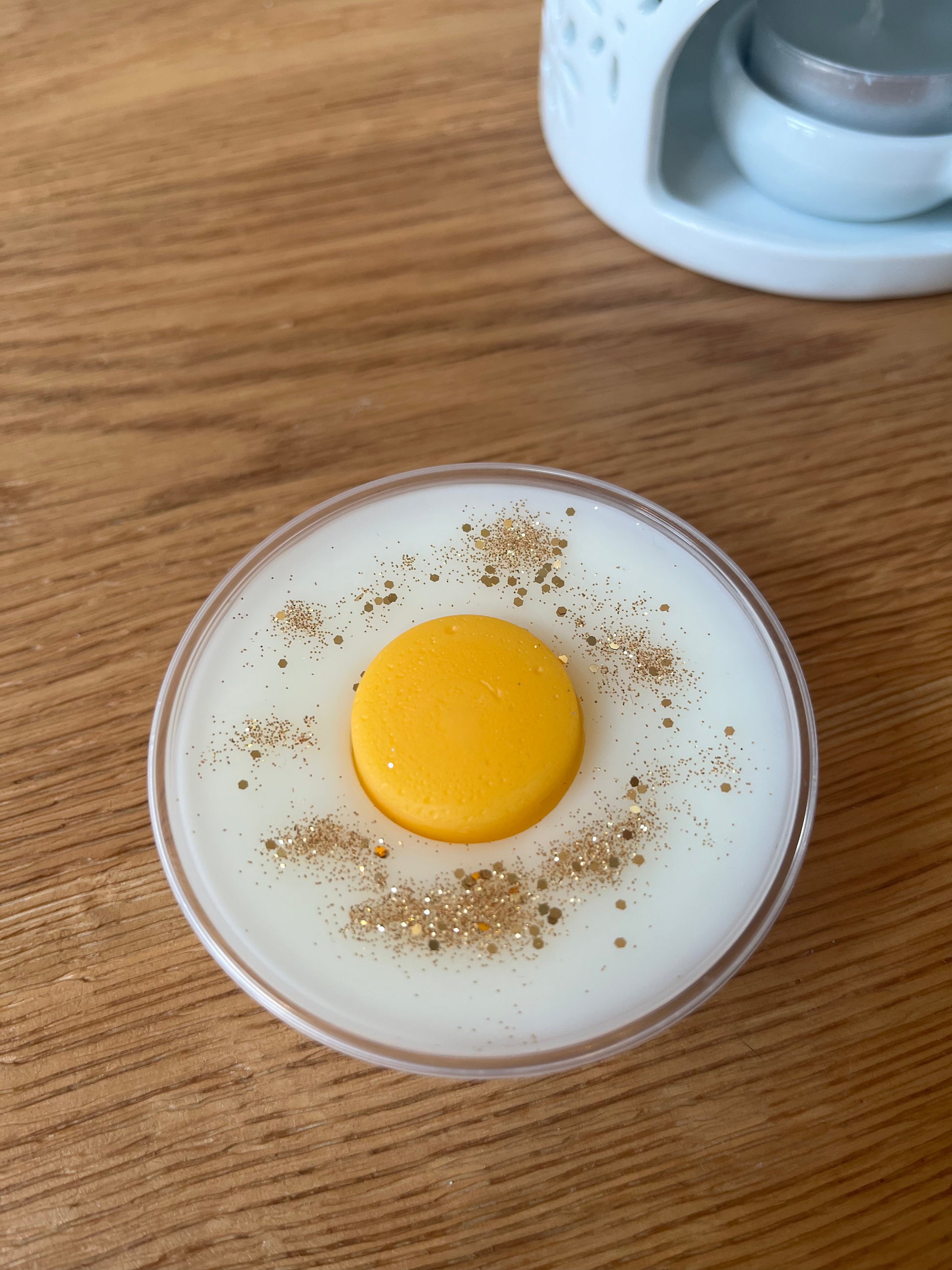 A pot of white wax topped with an orange wax disc and gold glitter with a wax melt burner in the background