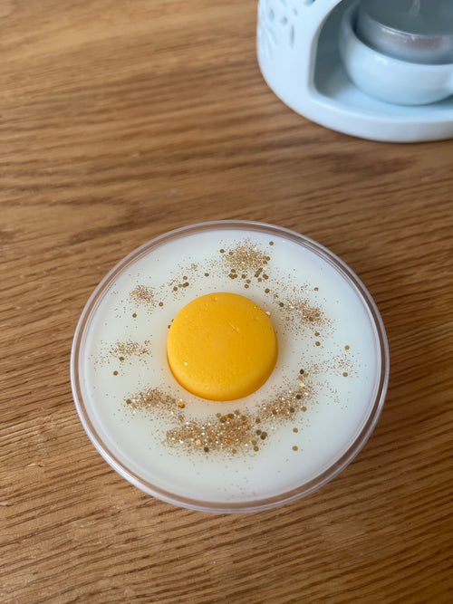 A pot of white wax topped with an orange wax disc and gold glitter with a wax melt burner in the background