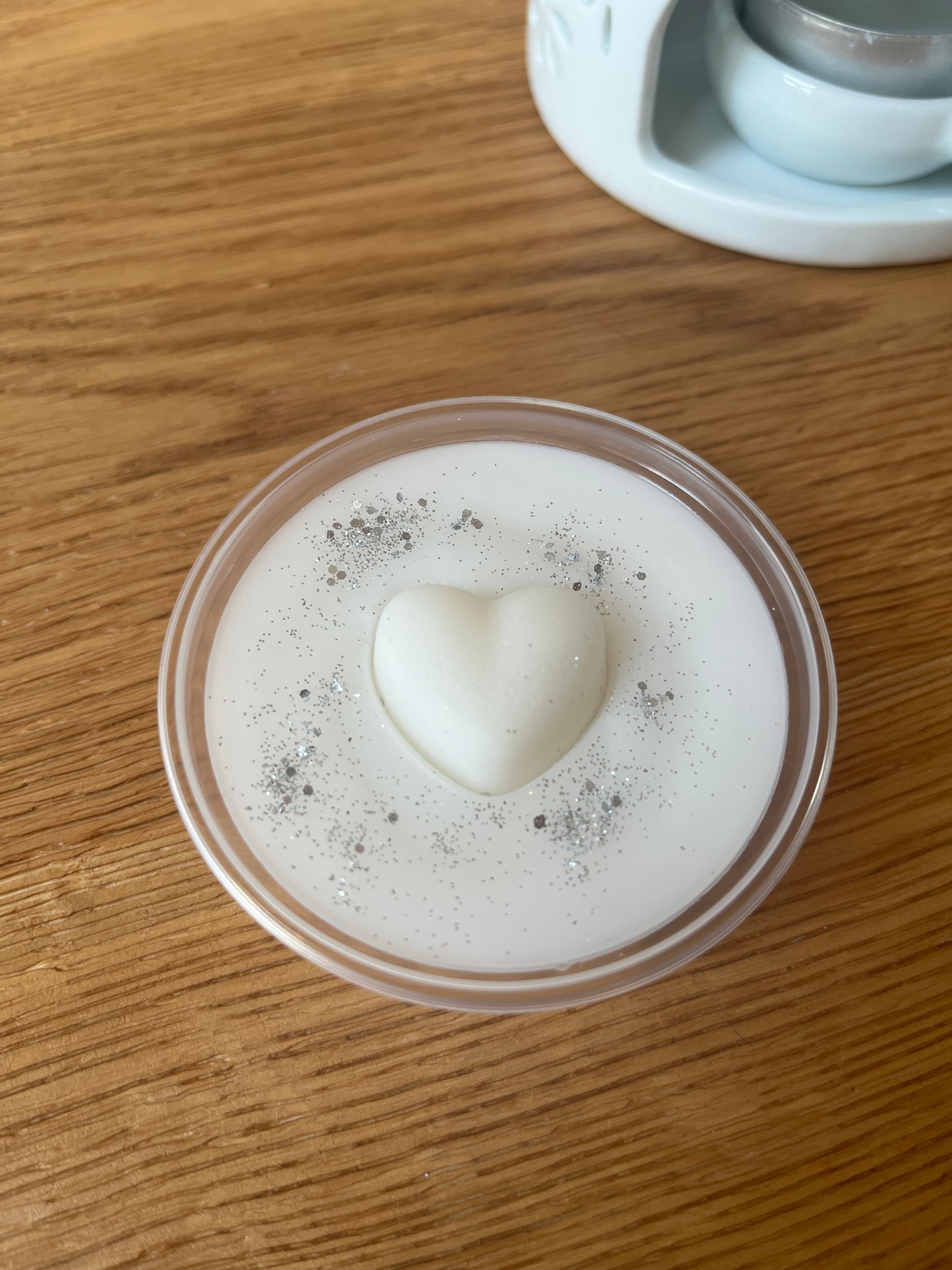 <img src="Fresh Linen_Pot" alt="Round container with a white wax melt featuring a heart-shaped top and silver glitter, placed on a wooden surface with part of a candle holder visible in the background.">