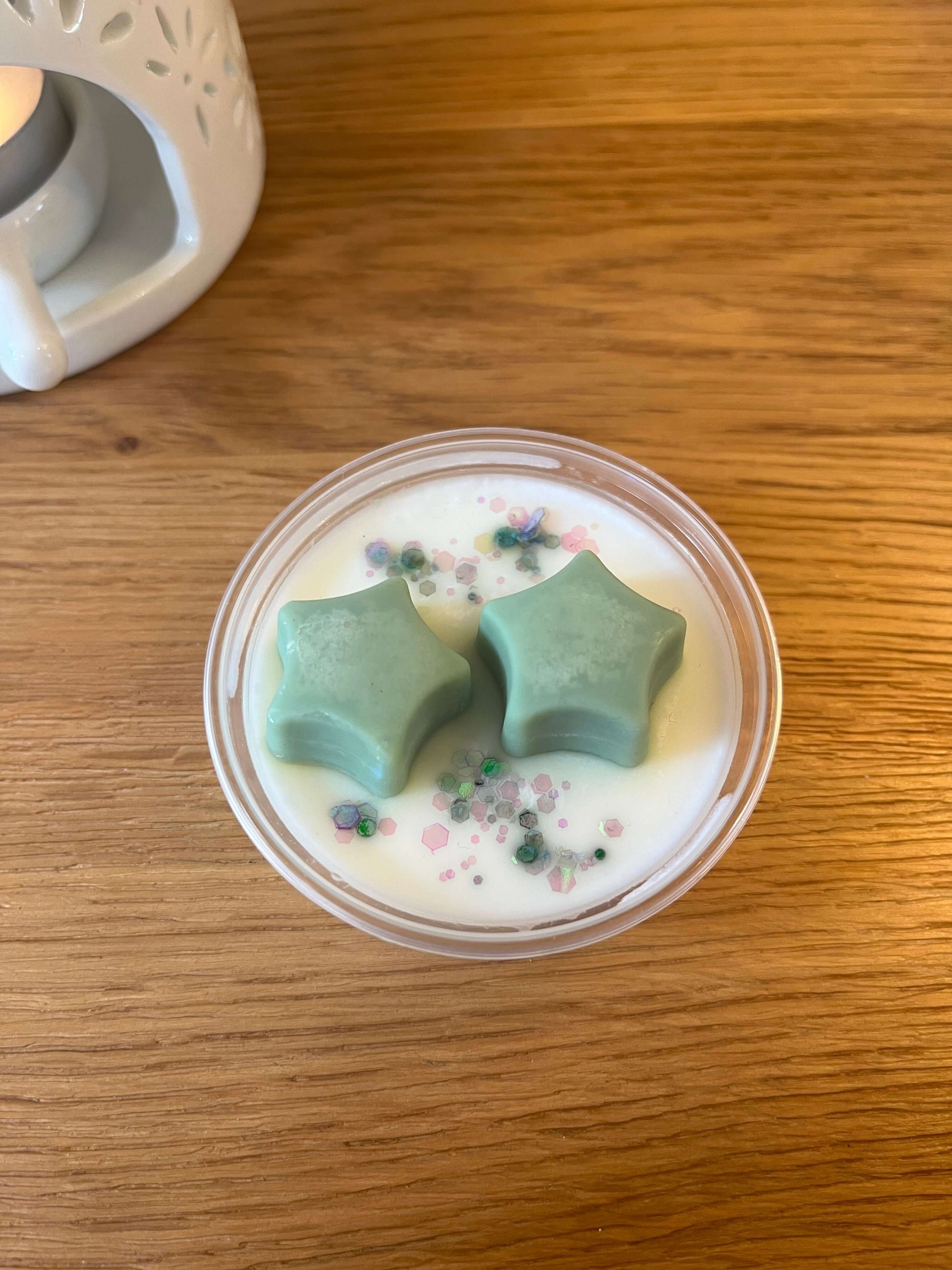 A pot of white wax topped with tow green stars and green glitter with a wax melt burner in the background