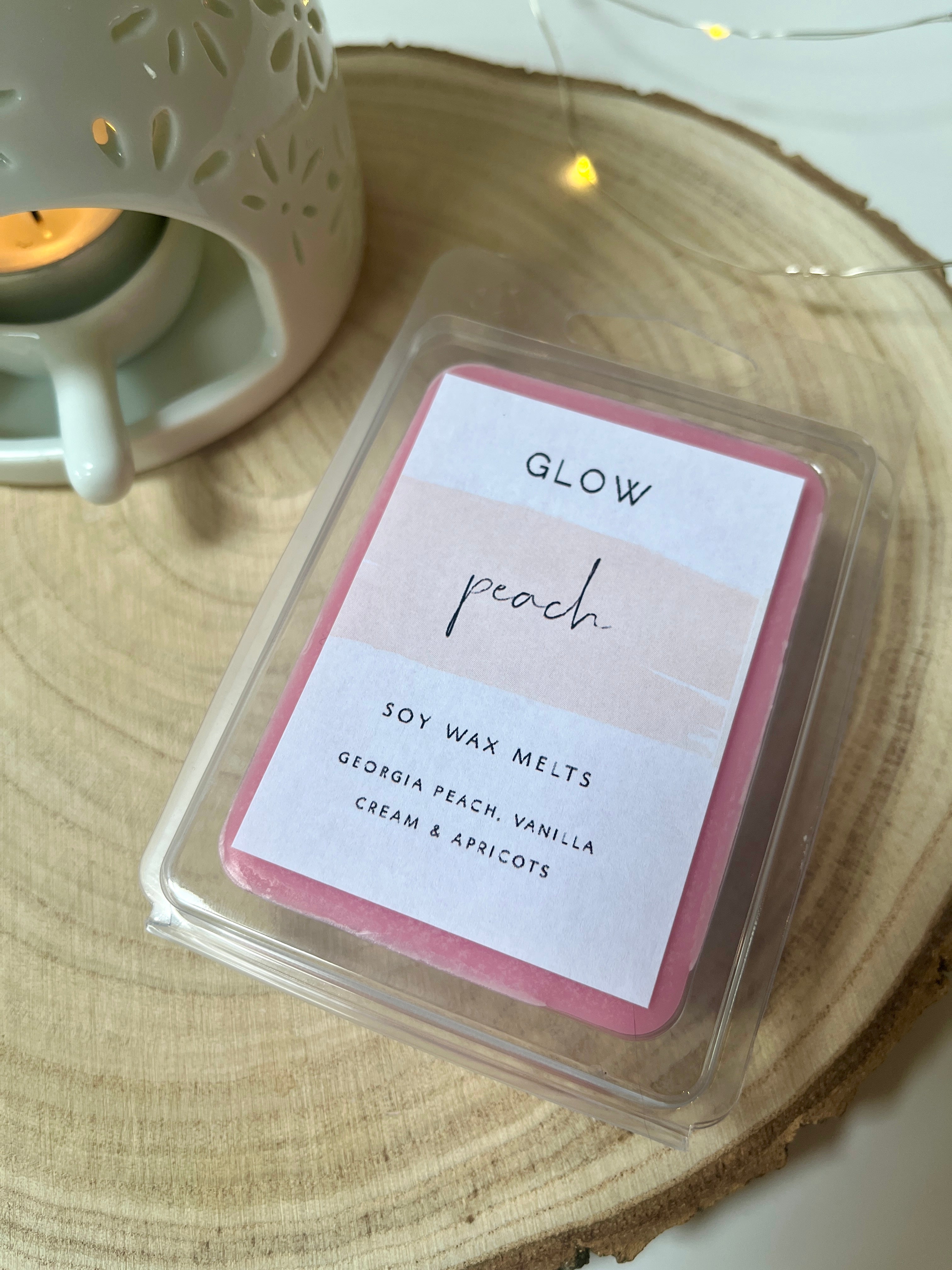 <img src="your-image-url.jpg" alt="Package of Glow soy wax melts labelled 'Peach' with scents of Georgia peach, vanilla, cream, and apricots, placed beside a white ceramic wax warmer with a lit candle.">