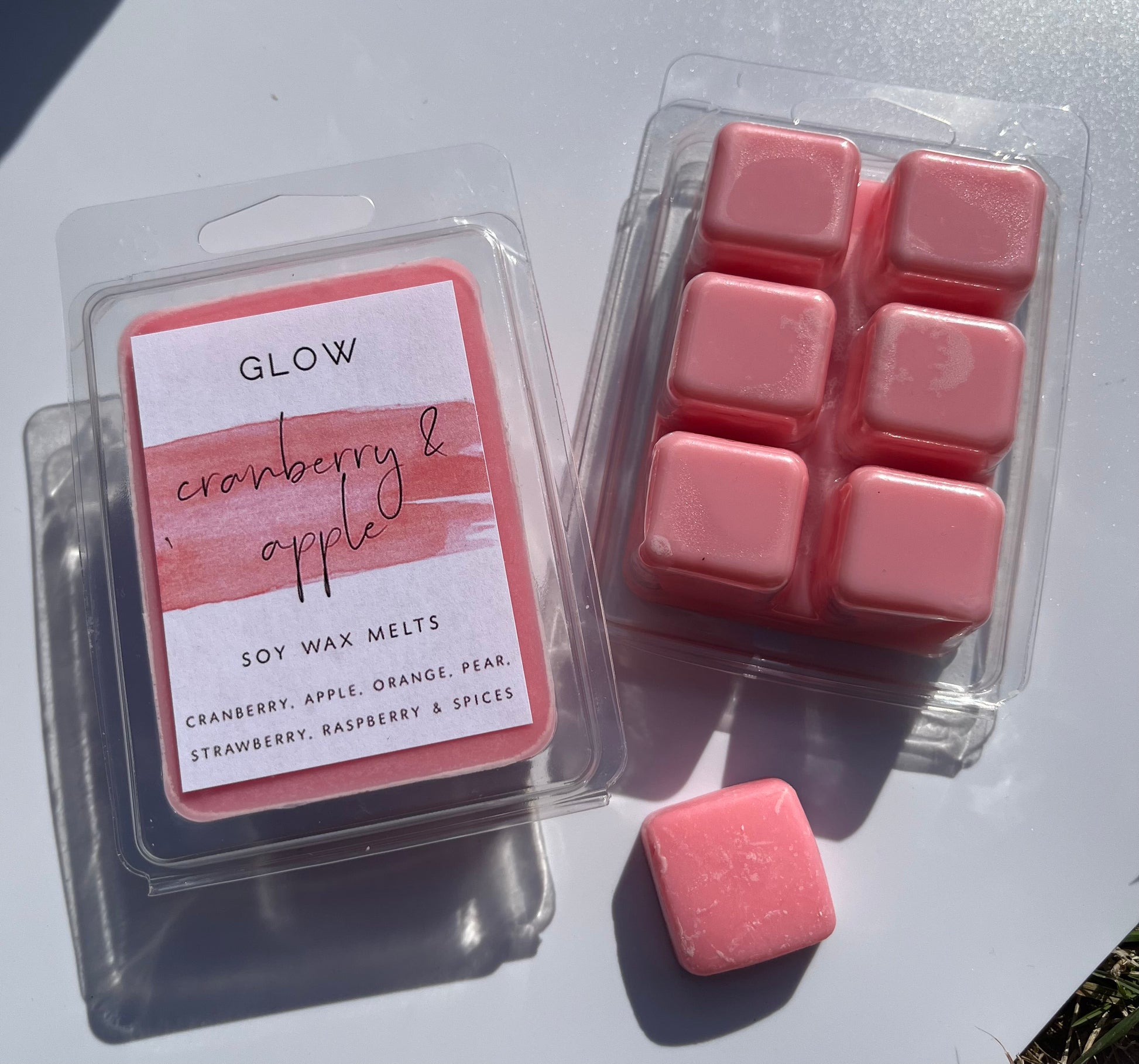 <img src="Cranberry Apple Clamshell" alt="Two packages of GLOW soy wax melts labelled 'Cranberry & Apple,' with six red square melts.">